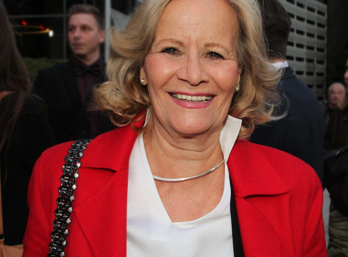 Uśmiechnięta kobieta w średnim wieku ubrana w czerwony płaszcz i białą bluzkę, stojąca wśród ludzi na tle nowoczesnego budynku.