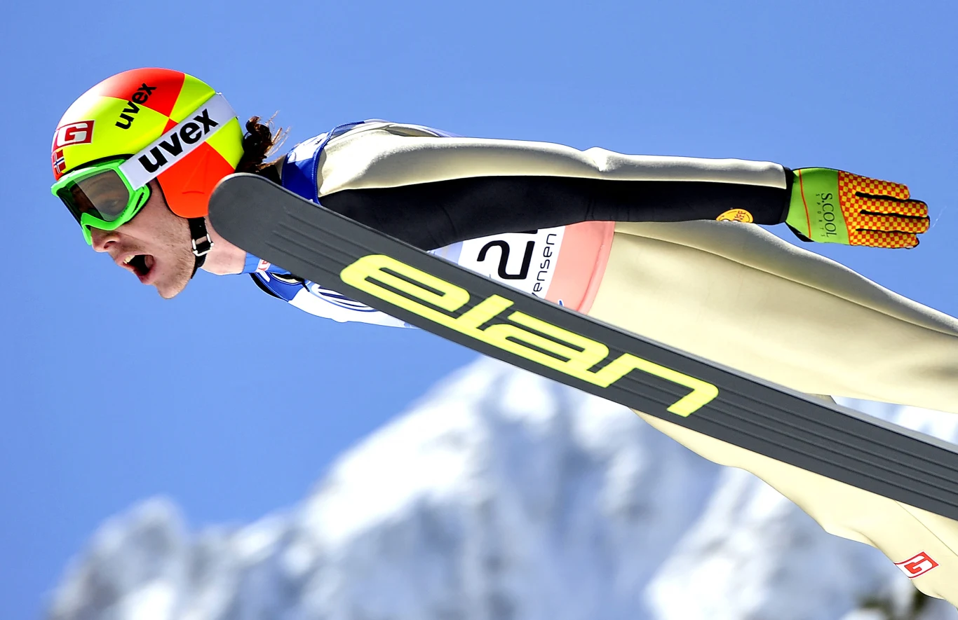 Zawodnik w stroju do skoków narciarskich wykonuje skok na tle ośnieżonych gór, widoczne narty marki Elan oraz kask ochronny.