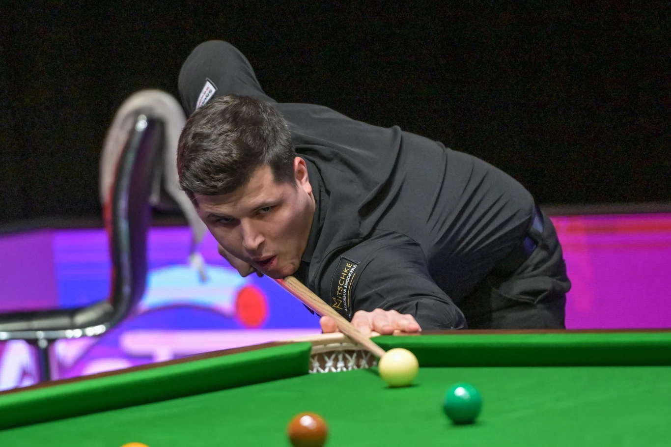 Mężczyzna w czarnym stroju skupiony podczas uderzenia bili na stole bilardowym, tło zabarwione różowym światłem, widoczne kule do gry.