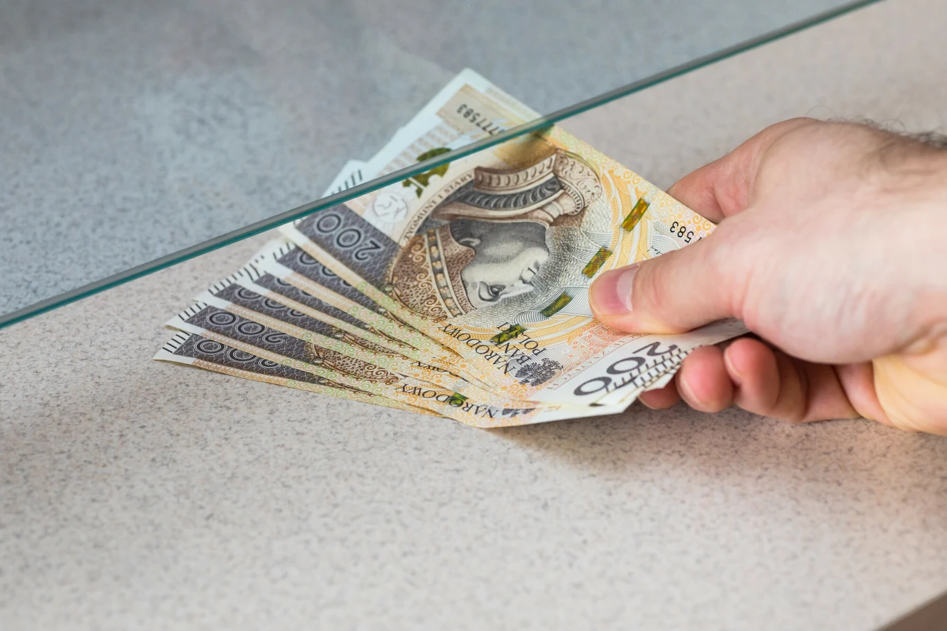 Banki ruszyły po klientów. Konkurencja rośnie, kredyt tanieje Ręka trzymająca kilka banknotów dwustuzłotowych, przekazująca je przez szybę, na tle jasnego blatu.
