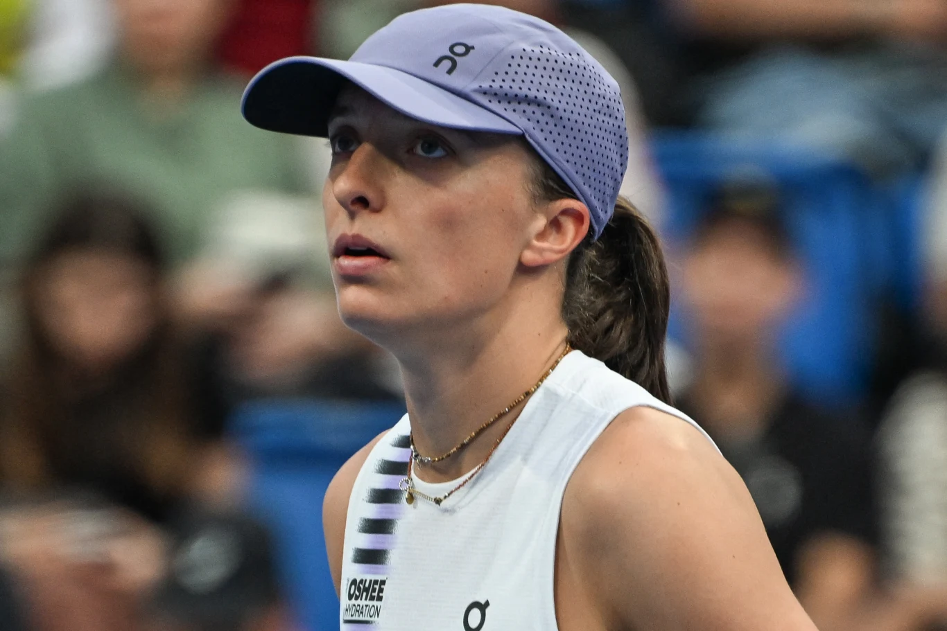 Młoda kobieta w sportowej czapce i tenisowym stroju patrząca w górę, na tle rozmytej publiczności.