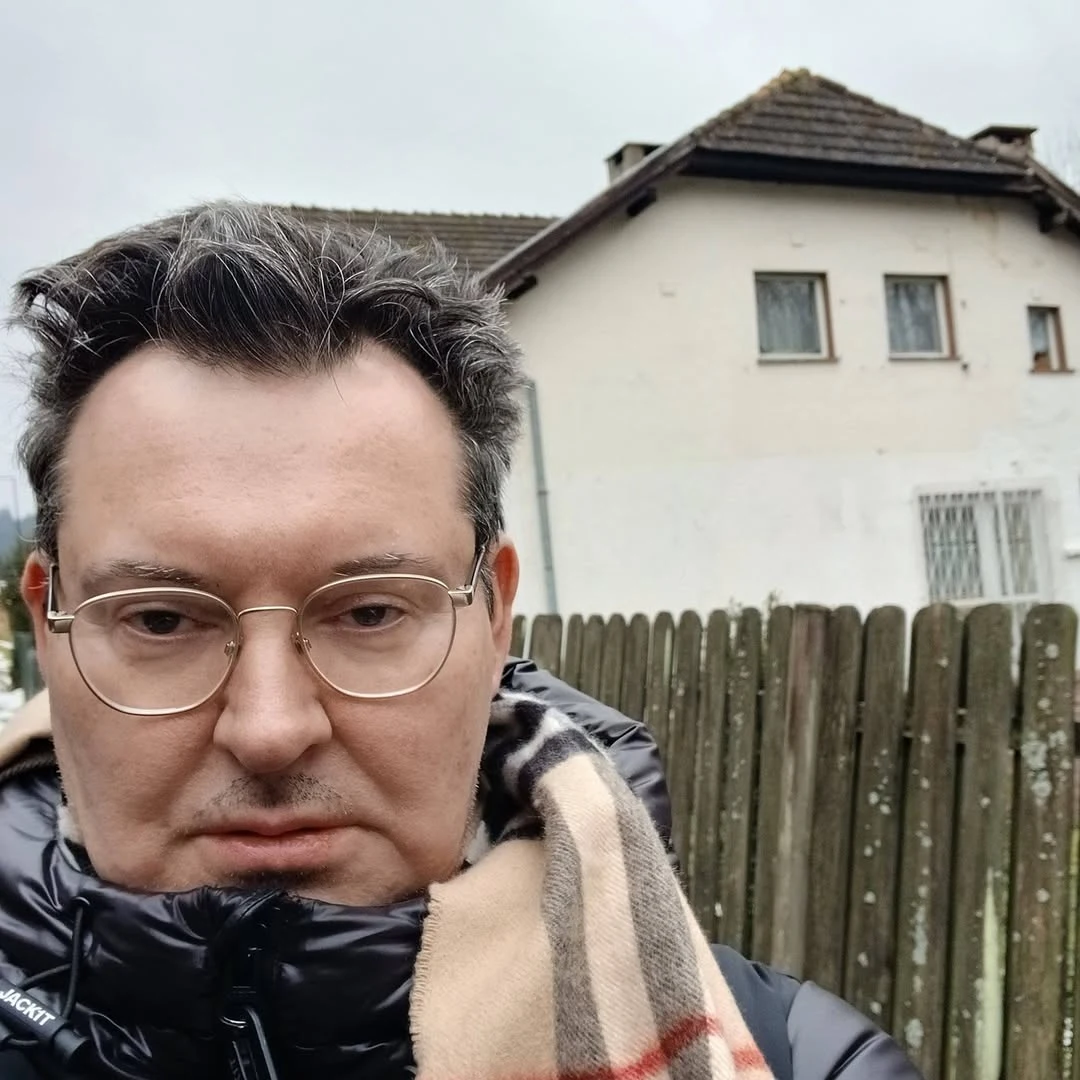 Mężczyzna w okularach i kurtce z apaszką stoi przed drewnianym płotem, w tle biały budynek mieszkalny o spadzistym dachu i zamkniętych oknach.