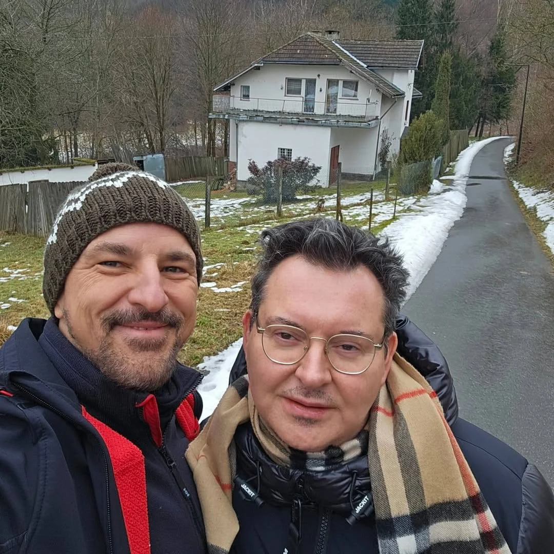 Dwóch mężczyzn pozuje razem do selfie na tle wiejskiej drogi oraz domu jednorodzinnego w zimowej scenerii, z otaczającym je sporadycznie śniegiem i drzewami.