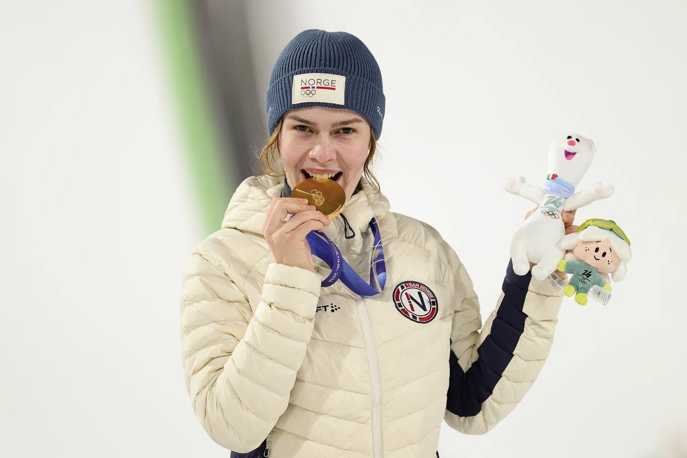 Sportsmenka w beżowej kurtce oraz niebieskiej czapce, gryzie złoty medal i trzyma w drugiej ręce maskotki. Za nią neutralne, rozmyte tło.