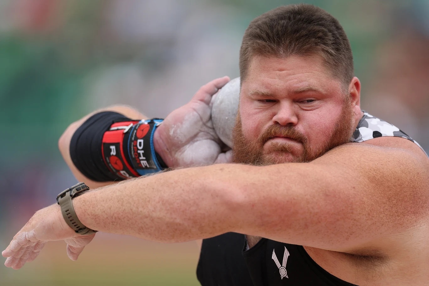 Mężczyzna o atletycznej budowie ciała przygotowuje się do pchnięcia kulą, skupiony, z widoczną kredą na dłoni oraz ochronną opaską na przedramieniu, w tle rozmyte trybuny stadionu sportowego.