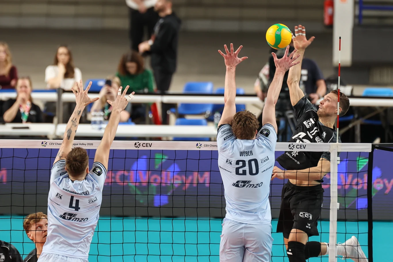 Dwóch siatkarzy drużyny w jasnych strojach próbuje zablokować atak zawodnika w czarnym stroju, który skacze i wykonuje atak nad siatką do piłki, widoczna publiczność na trybunach.