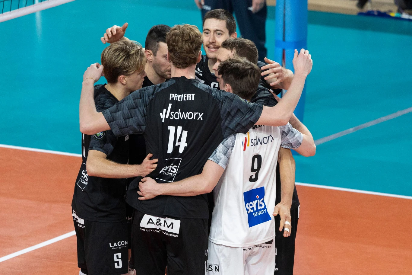 Grupa siatkarzy w czarno-białych strojach świętuje punkt, obejmując się na boisku tuż przy siatce sportowej.