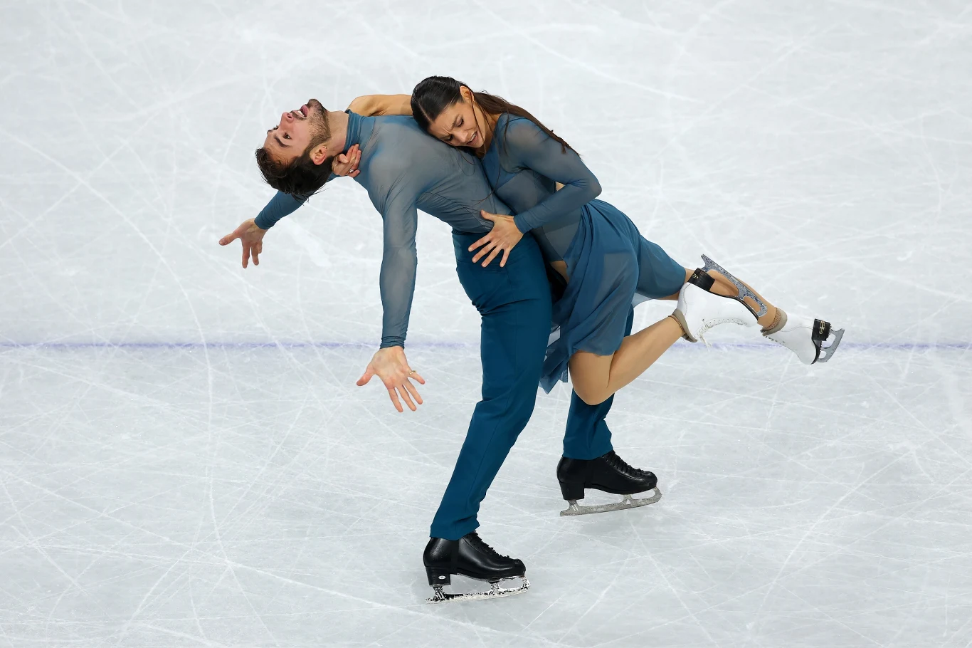 Para łyżwiarzy figurowych wykonuje dynamiczną figurę na lodowisku, oboje ubrani w dopasowane niebieskie kostiumy, wyrażając emocje i harmonię ruchu.