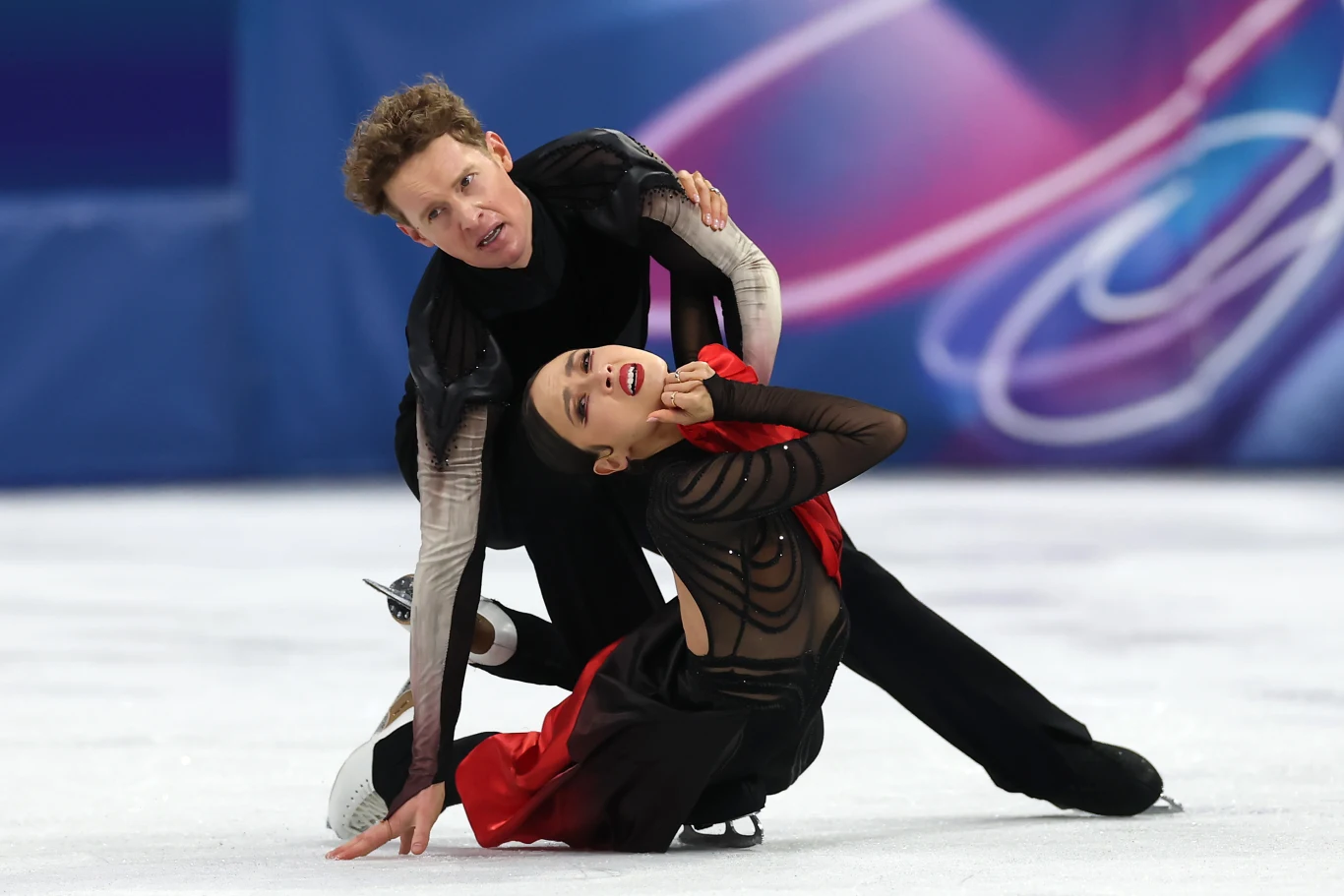 Para łyżwiarzy figurowych podczas widowiskowego elementu tanecznego na lodzie, kobieta w eleganckiej czarno-czerwonej sukni, partner w czarnym kostiumie, dynamiczne pozy, kolorowe tło z rozmytymi wzorami.