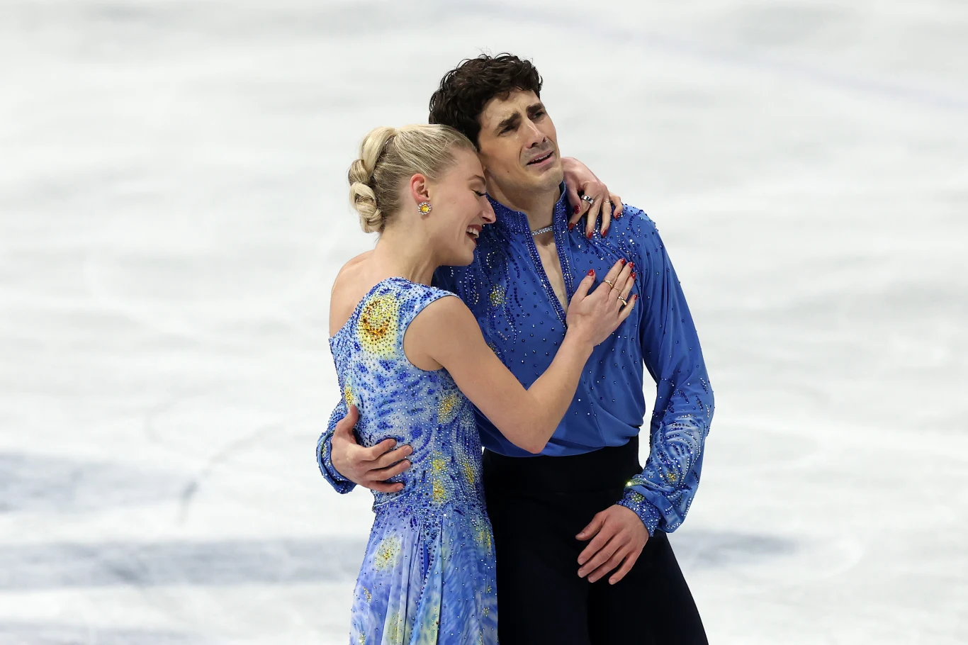 Para łyżwiarzy figurowych obejmuje się na lodowisku, ubrani są w niebieskie stroje sceniczne, wokół nich rozciąga się gładka tafla lodu.