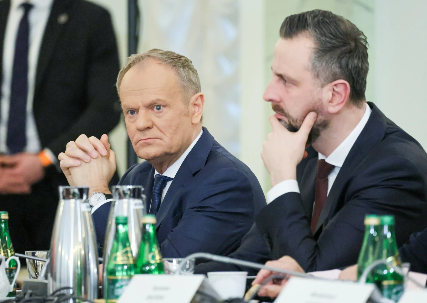 Premier Donald Tusk oraz wicepremier i szef MON Władysław Kosiniak-Kamysz Dwóch mężczyzn w garniturach siedzi przy stole konferencyjnym, skupieni i poważni, wokół butelki wody oraz metalowy dzbanek, w tle widać innych uczestników spotkania.