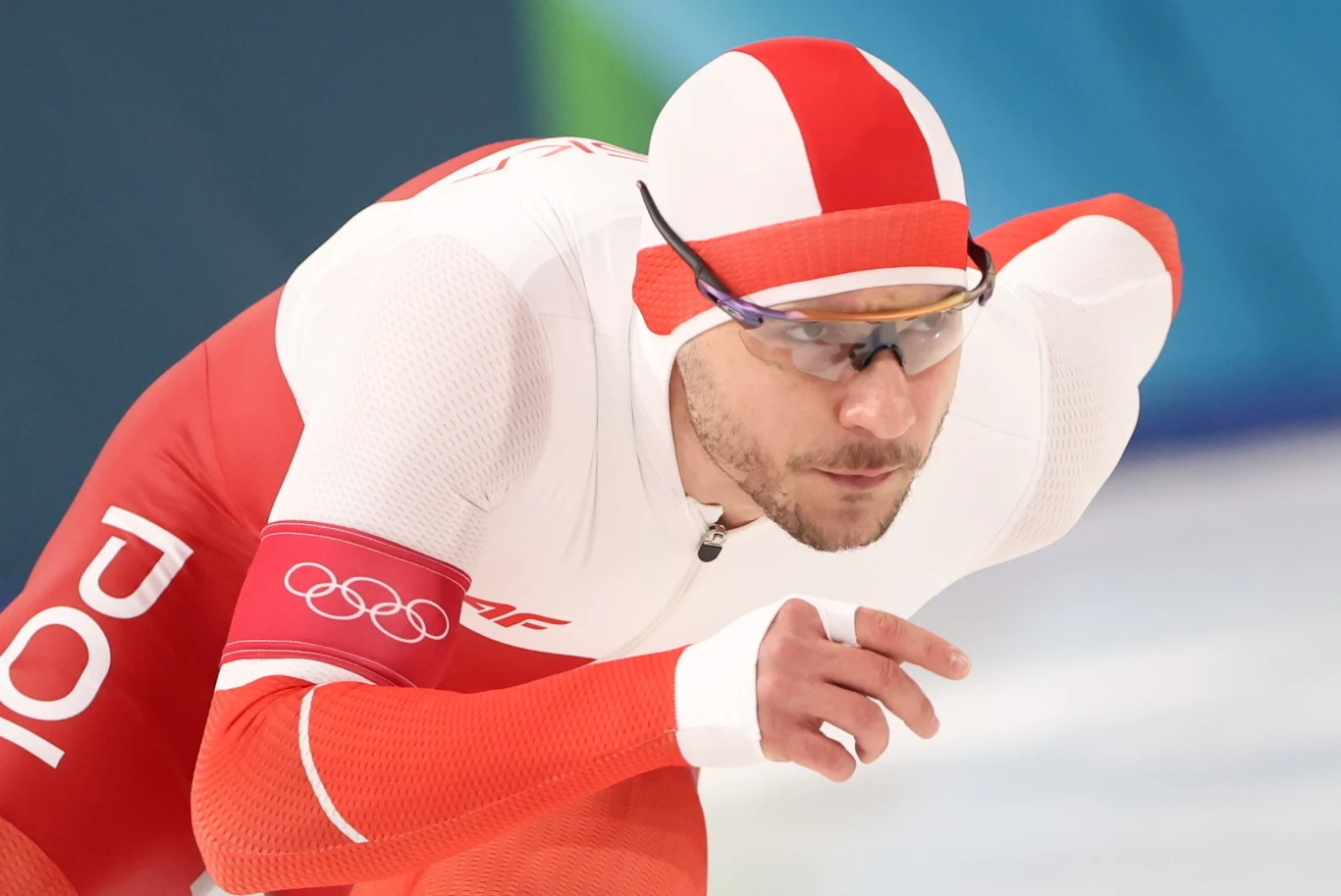Zawodnik w stroju panczenisty w barwach Polski podczas wyścigu na łyżwach szybkich na lodowisku, skoncentrowany w pozycji startowej, z opaską olimpijską na ramieniu.
