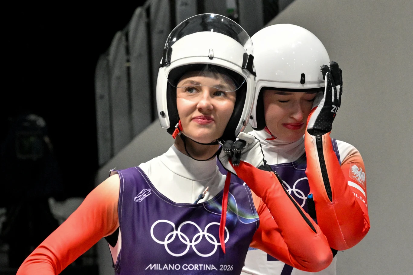 Dominika Piwkowska (z przodu) i Nikola Domowicz Dwie zawodniczki w kombinezonach i kaskach ochronnych stoją na torze podczas zimowych igrzysk olimpijskich, na ich strojach widnieje logo igrzysk Milano Cortina 2026.