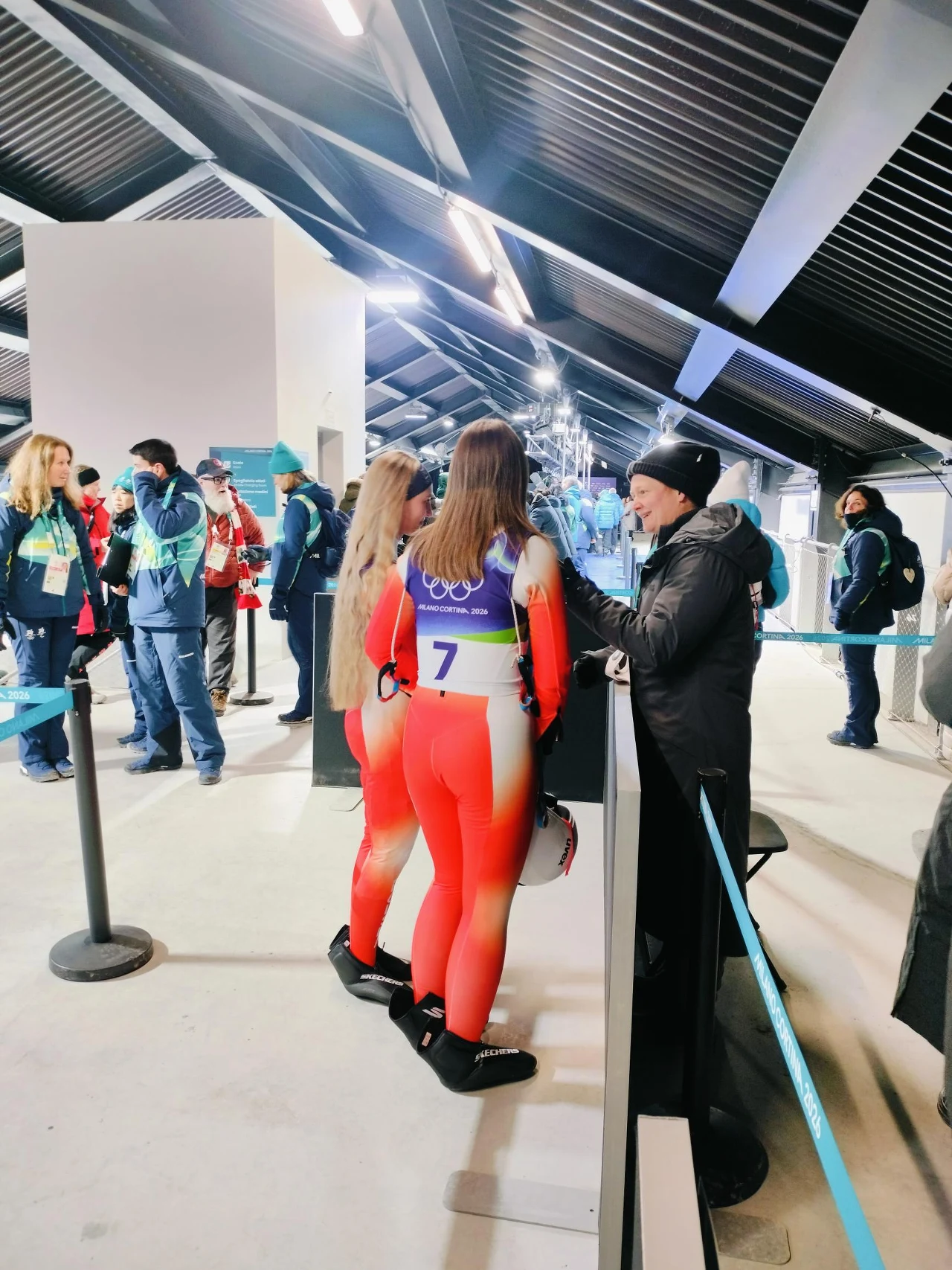 Zawodnicy w kombinezonach sportowych, z numerem startowym 7, stoją w kolejce do stanowiska obsługi, otoczeni innymi uczestnikami oraz personelem w odzieży zimowej, wnętrze budynku o nowoczesnej konstrukcji z dachem z blachy.