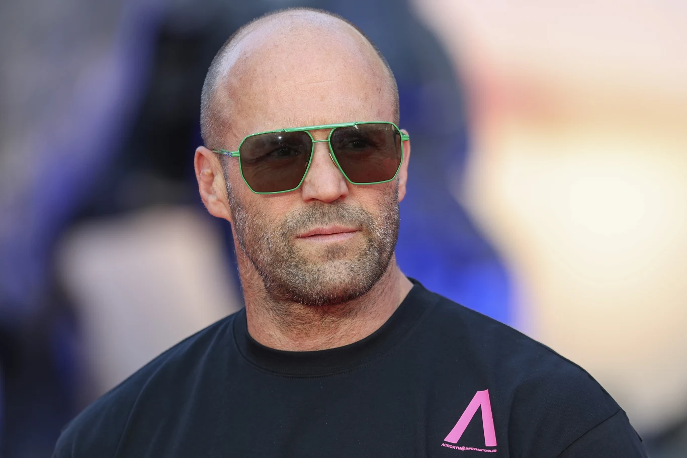 Jason Statham / WireImage Łysy mężczyzna w zielonych okularach przeciwsłonecznych i czarnej koszulce z różowym logo patrzy poważnie w dal, tło rozmyte w niebieskich odcieniach.