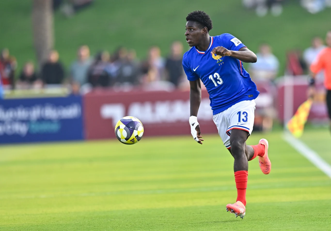 Piłkarz w niebieskiej koszulce z numerem 13 biegnie po boisku, koncentrując się na lecącą piłkę. W tle rozmazani kibice i zielona murawa.