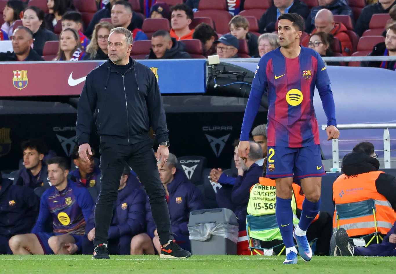 Trener i piłkarz stoją przy linii bocznej boiska na tle ławki rezerwowych oraz kibiców na trybunach, piłkarz ubrany w koszulkę FC Barcelony z numerem 2, atmosfera meczu piłkarskiego.