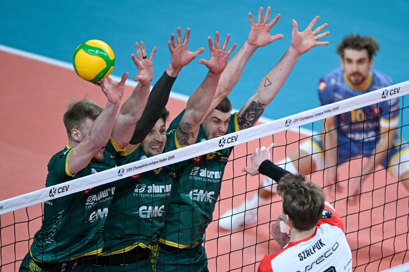 Atak Artura Szalpuka w meczu z Aluron CMC Wartą Zawiercie Trzech siatkarzy w zielonych koszulkach wykonuje blok przy siatce, próbując zatrzymać atakującego zawodnika drużyny przeciwnej w białej i czerwonej koszulce z nazwiskiem 'Szum' na plecach, w tle widoczny jest kolejny zawodnik obserwujący sytuację.