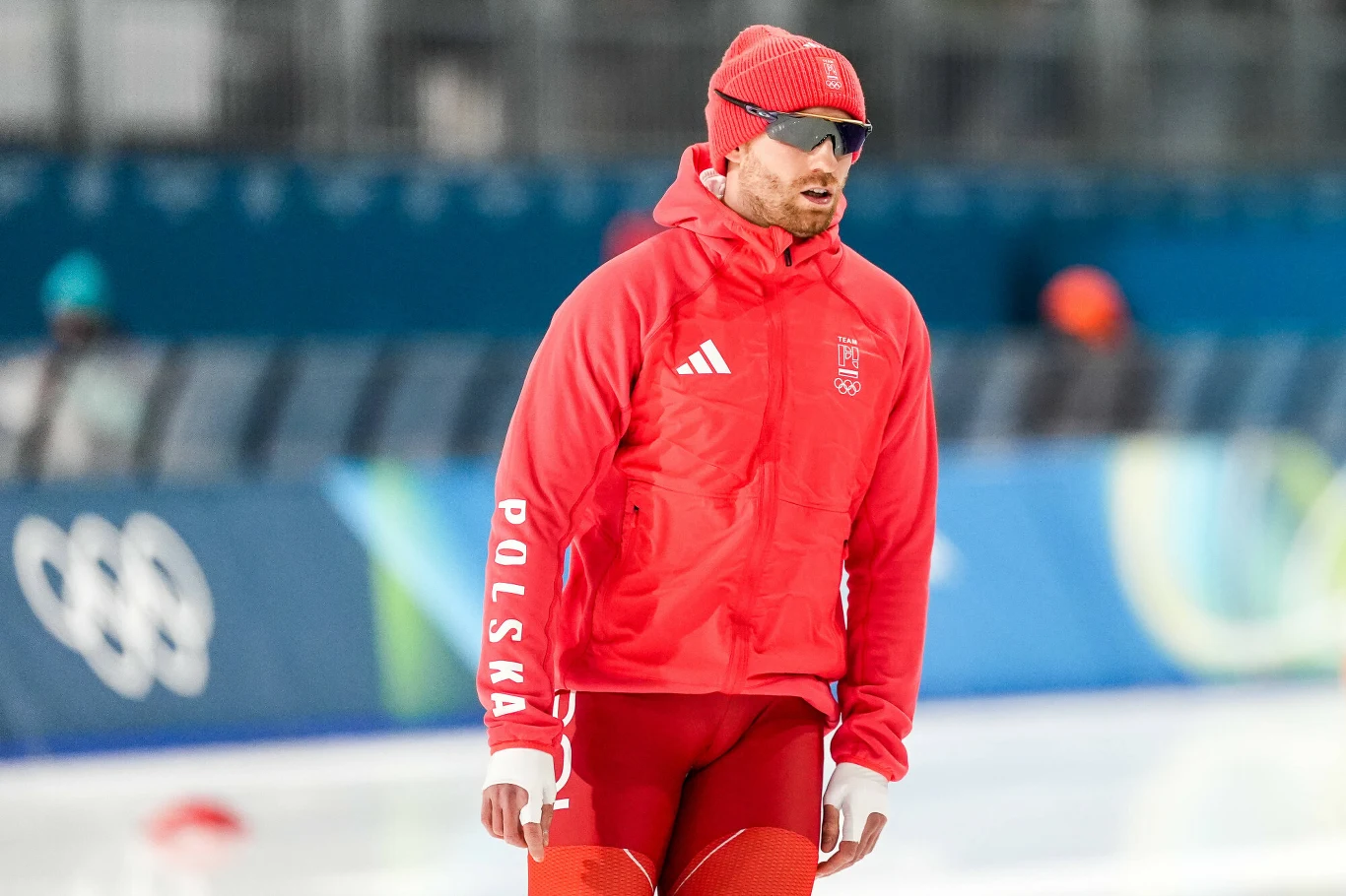 Damian Żurek Zawodnik ubrany w czerwoną zimową odzież sportową z napisem 'Polska', stojący na lodowisku, w tle widoczne rozmyte elementy stadionu olimpijskiego.