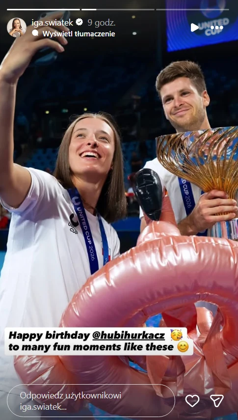 Dwoje uśmiechniętych sportowców pozuje razem na tle areny, trzymając puchar oraz dużą, dmuchaną różową dekorację w kształcie flaminga.