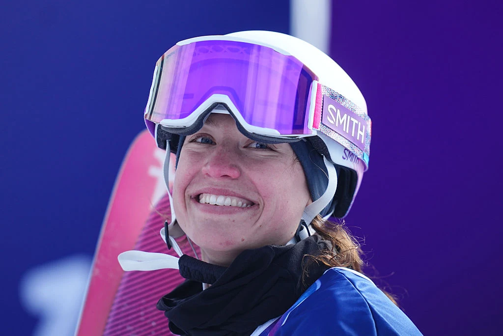 Uśmiechnięta kobieta w sportowym stroju narciarskim, z kaskiem i dużymi goglami, trzymająca snowboard na tle fioletowego rozmycia.