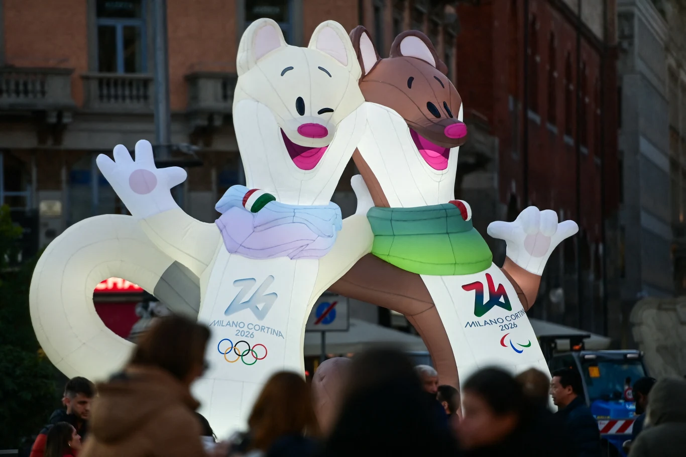 Gigantyczne dmuchane maskotki w kształcie zwierząt z logo Zimowych Igrzysk Olimpijskich Milano Cortina 2026 ustawione na tle miejskich budynków, otoczone tłumem ludzi.