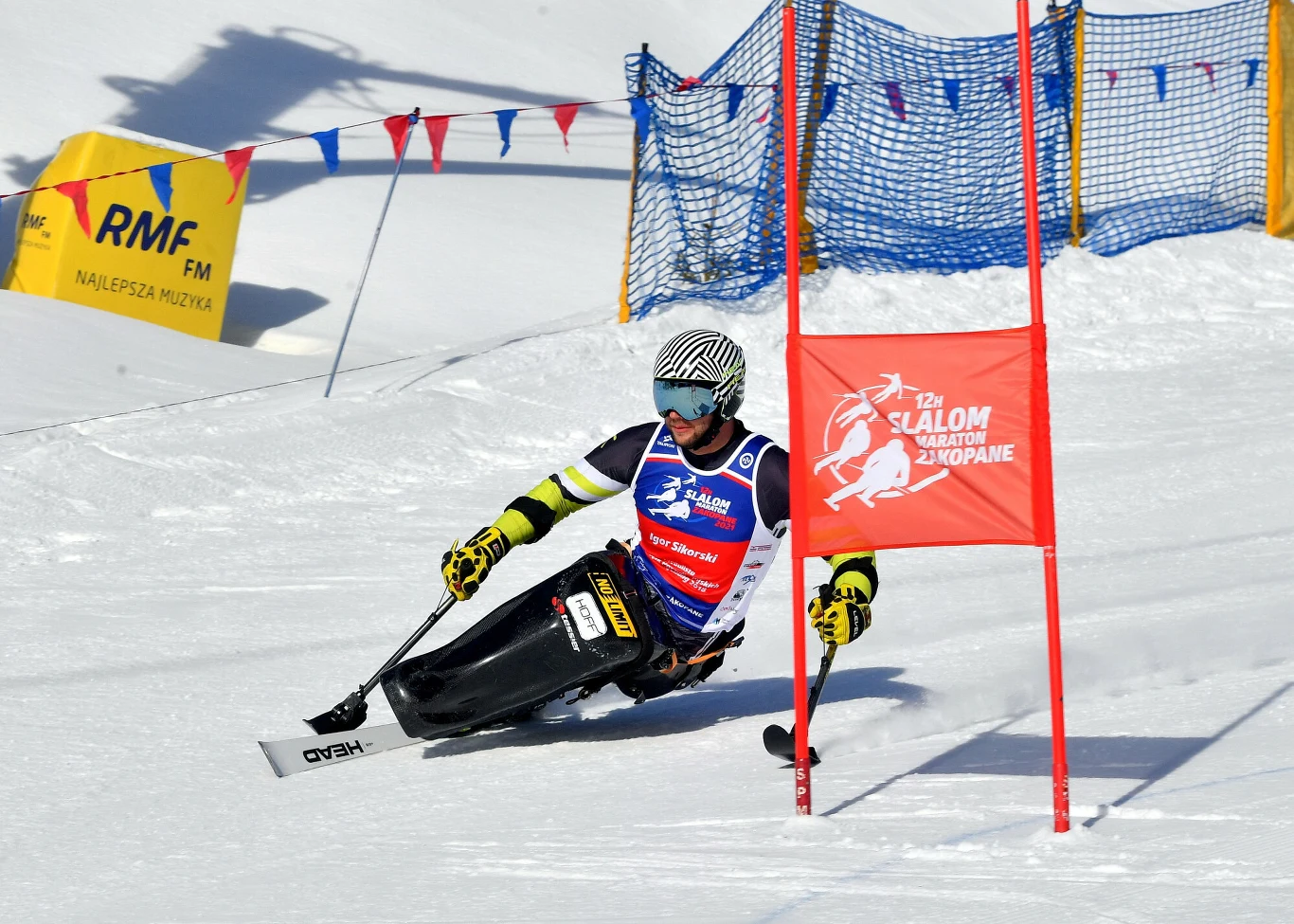 Zawodnik narciarstwa paraalpejskiego ściga się na specjalnym wózku monoski, pokonując zakręt między tyczkami na trasie slalomu giganta w zimowej scenerii, w tle widoczne bandy zabezpieczające i kolorowe chorągiewki.