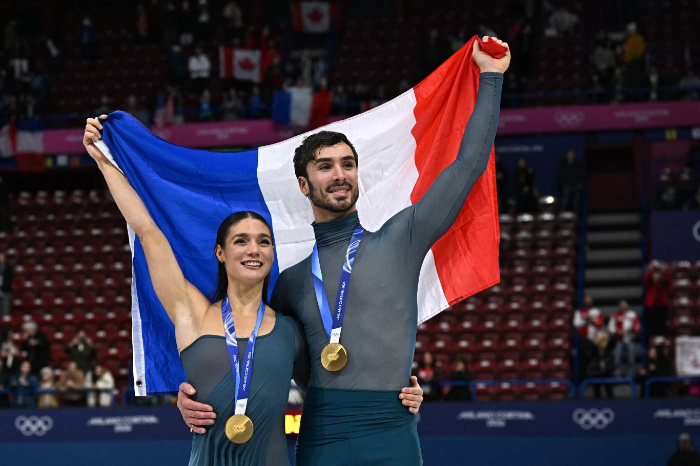 Laurence Fournier Beaudry i Guillaume Cizeron Dwoje sportowców w strojach do łyżwiarstwa figurowego trzyma francuską flagę, na szyjach mają złote medale, w tle trybuny i wywieszone flagi różnych państw.