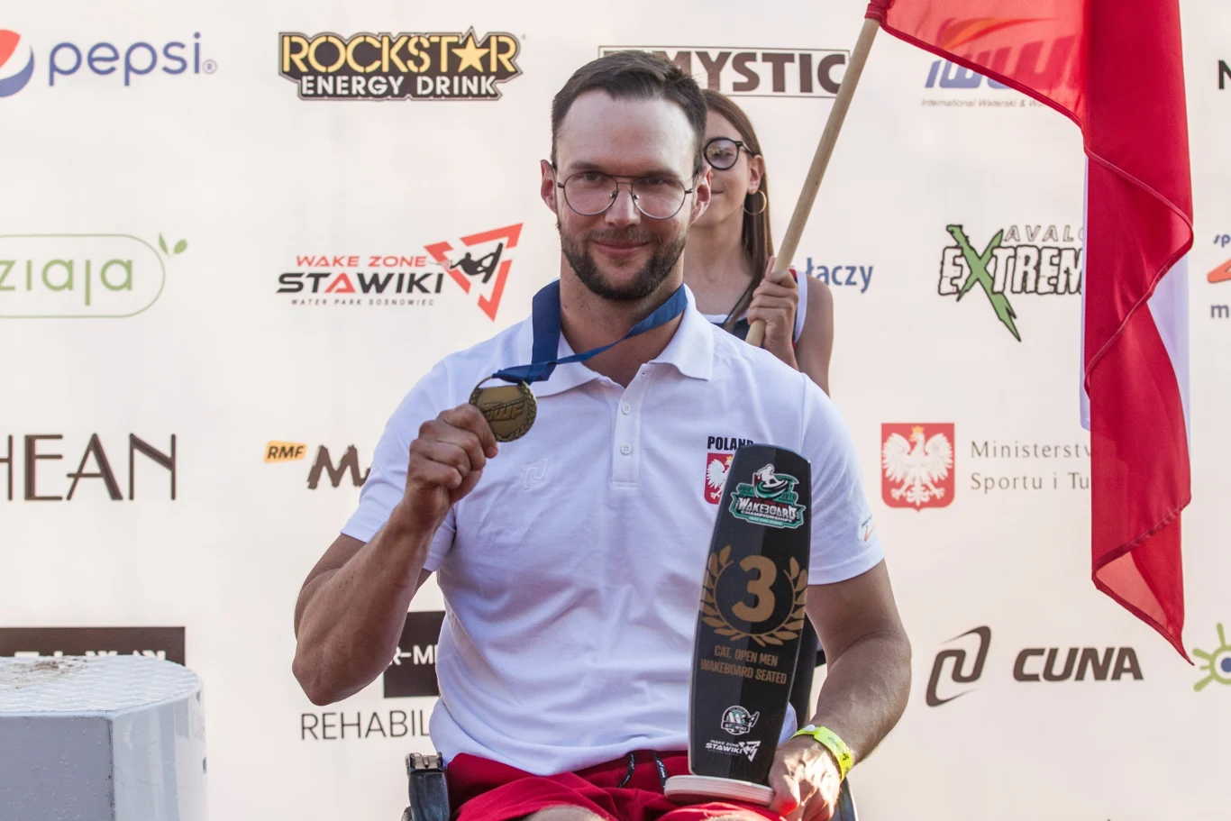 Mężczyzna na wózku inwalidzkim z medalem i pucharem za trzecie miejsce, ubrany w biały polos z godłem Polski, trzymający flagę, w tle widoczne loga sponsorów.