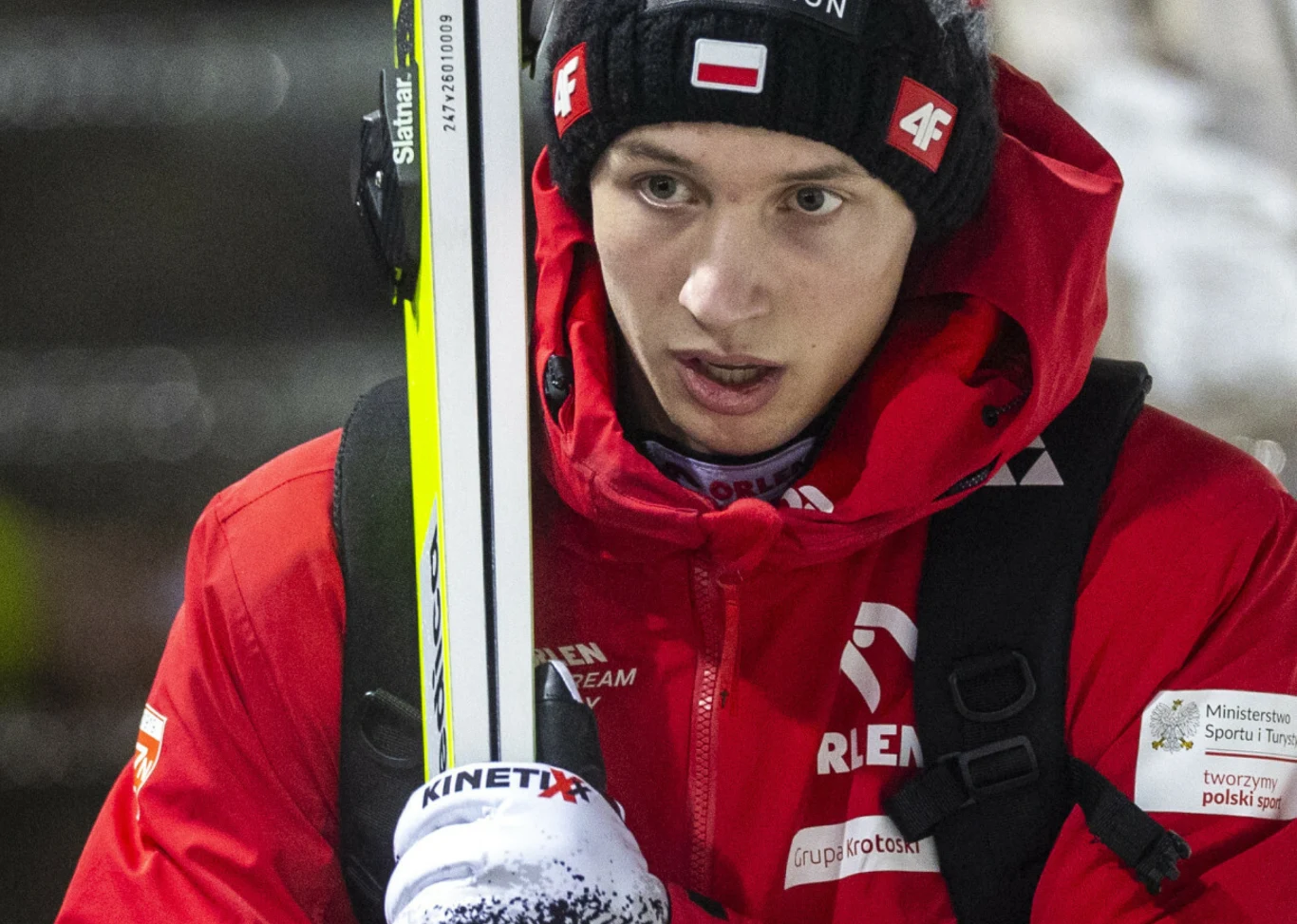 Na zdjęciu polski skoczek narciarski Kacper Tomasiak Młody narciarz w czerwonej kurtce sportowej z nartą wspartą na ramieniu, ubrany w zimową czapkę z polską flagą i logo sponsora, widoczne napisy na kurtce związane z polską reprezentacją