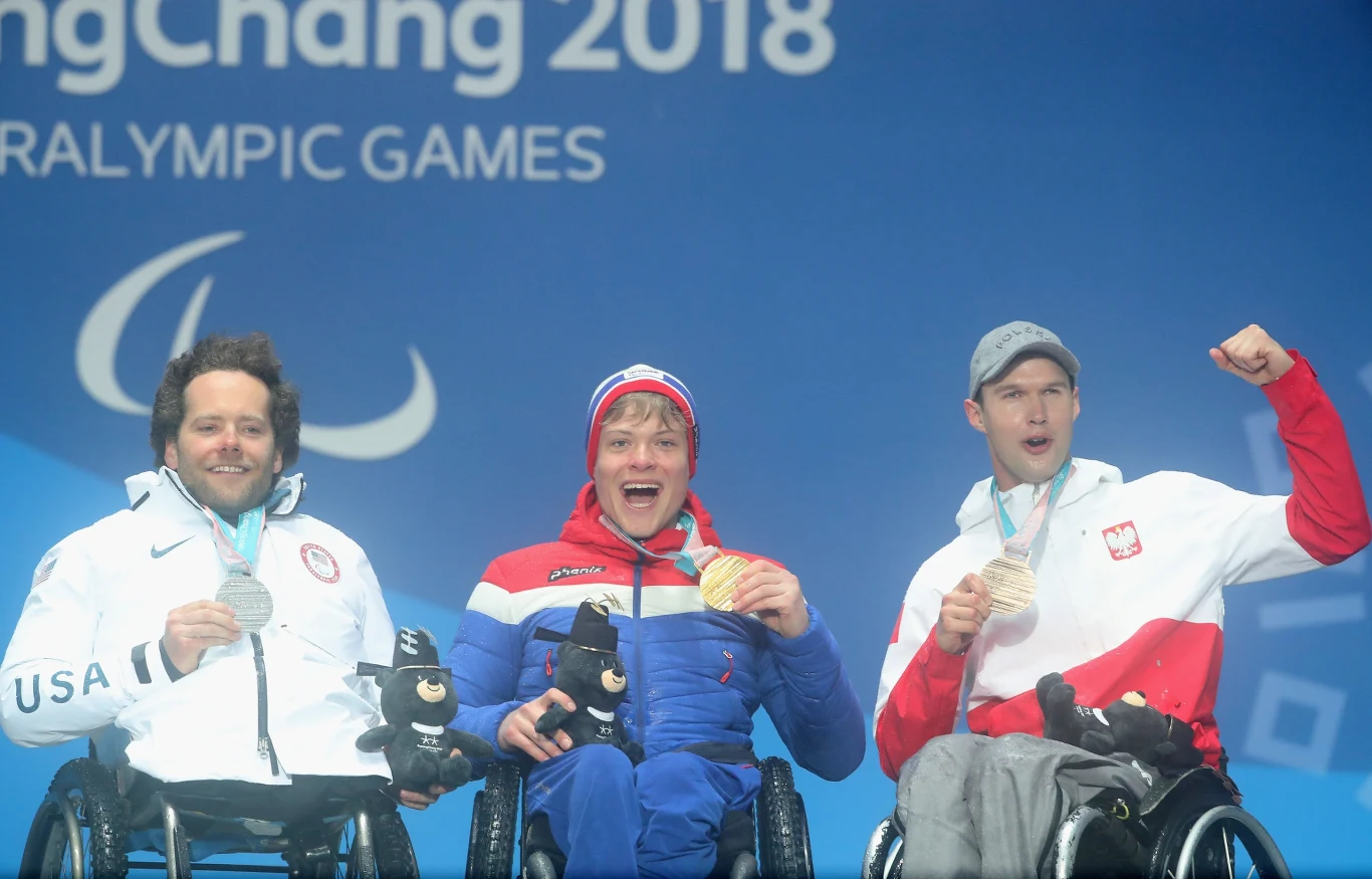 Trzech sportowców na wózkach inwalidzkich pozuje na podium, każdy z medalem oraz maskotką, ubrani w stroje reprezentacyjne, radośni i dumni z osiągnięć, w tle napis PyeongChang 2018 oraz logo igrzysk paraolimpijskich.