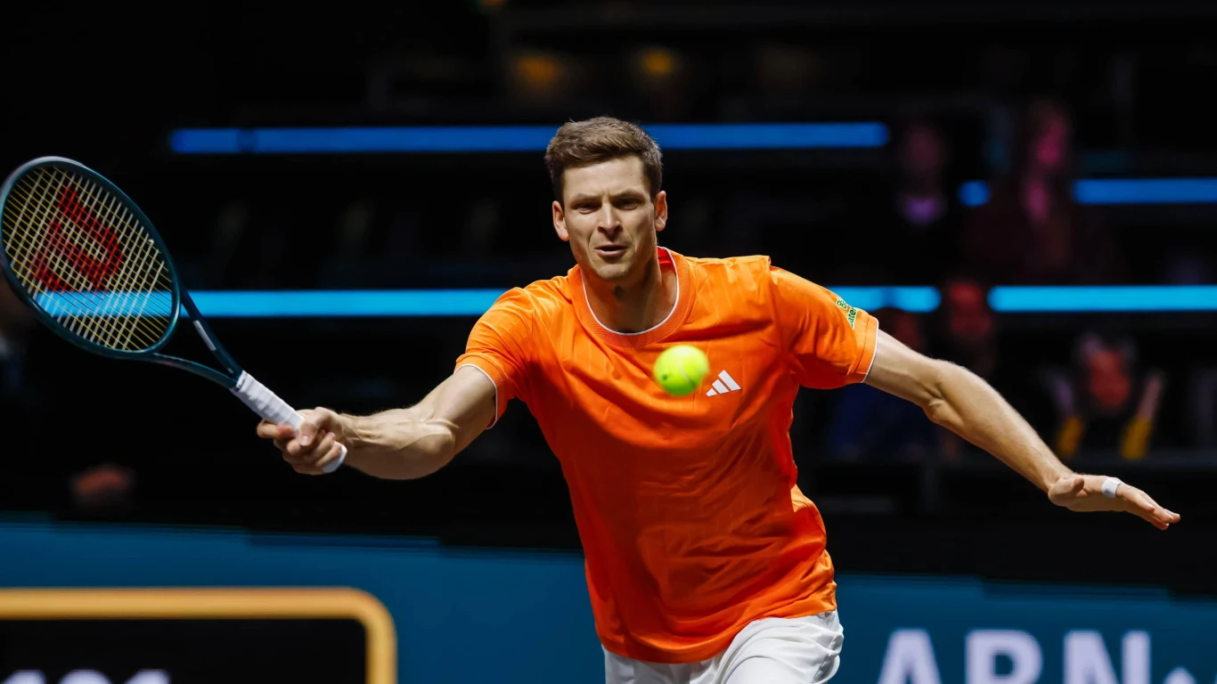 Tenisista w pomarańczowej koszulce skupiony na uderzeniu piłki podczas meczu, dynamiczna poza na tle ciemnej hali sportowej.