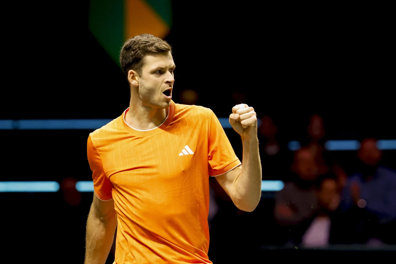 Mężczyzna w pomarańczowej koszulce sportowej pokazuje gest zaciśniętej pięści, wyrażając emocje podczas meczu tenisowego. W tle rozmazana publiczność oraz ciemna sceneria kortu.