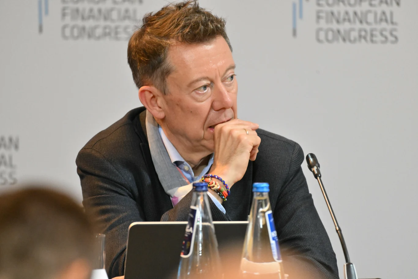 Przemek Gdanski, Prezes Zarzadu, BNP Paribas Bank Polska S.A. Mężczyzna siedzący przy stole konferencyjnym, zamyślony, opierający brodę na zaciśniętej dłoni, wokół dokumenty, laptop oraz butelki z wodą, w tle widoczne logo European Financial Congress.