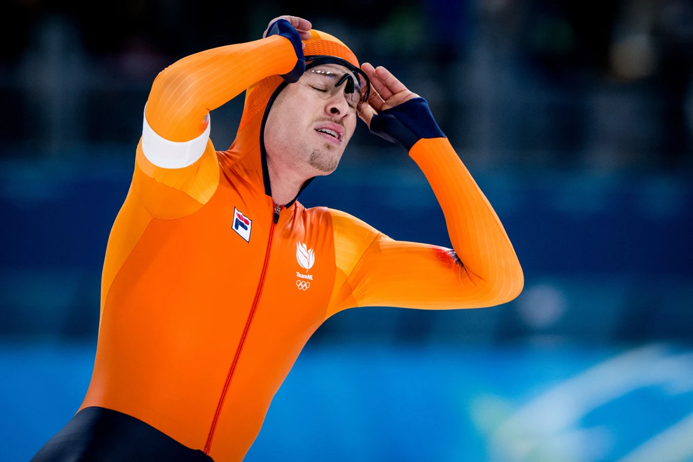 Joep Wennemars Łyżwiarz szybki w pomarańczowym stroju sportowym na tle lodowiska, trzymający dłonie na głowie, wyrażający silne emocje.