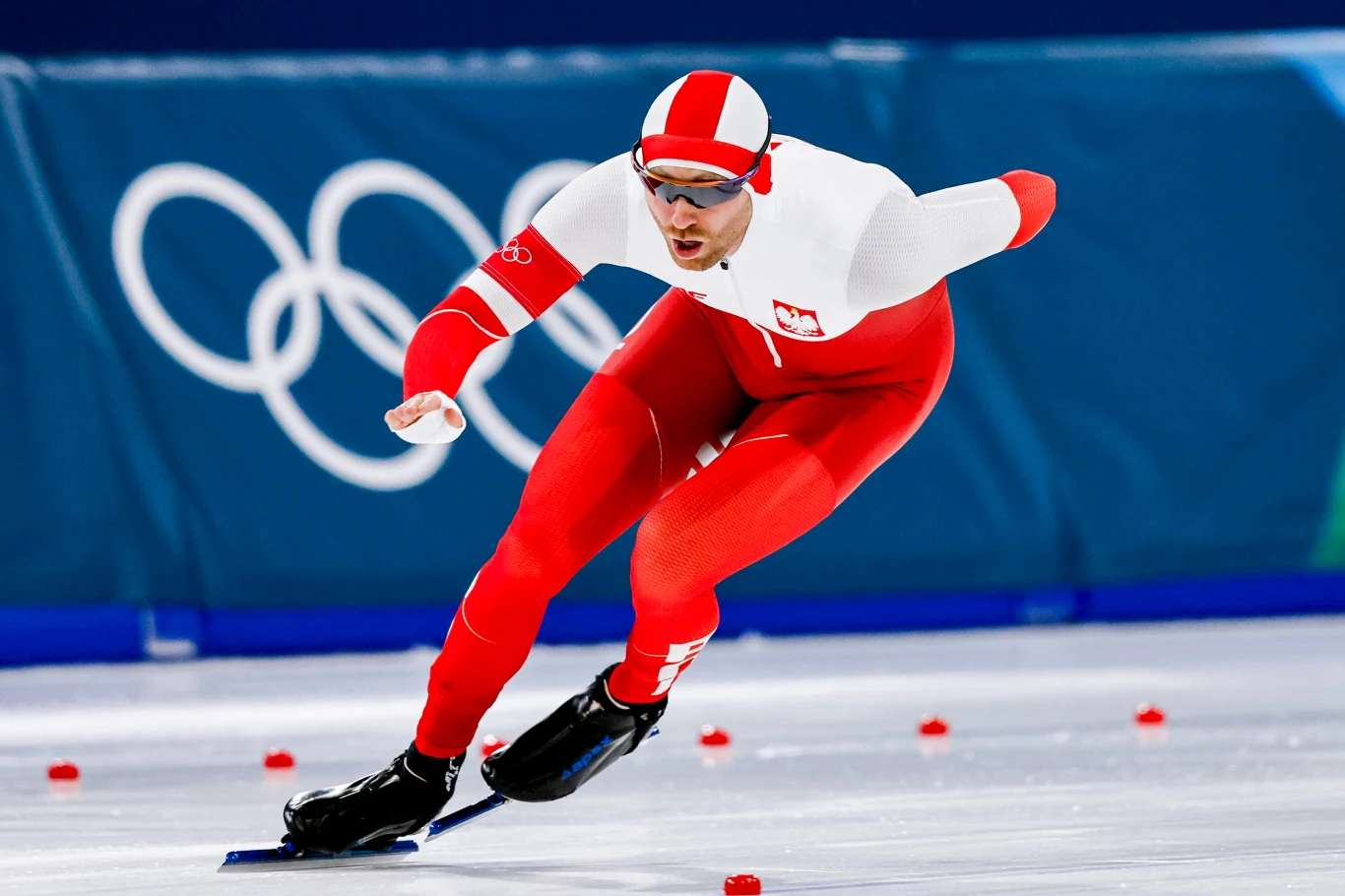 Damian Żurek Łyżwiarz szybki w biało-czerwonym stroju reprezentacji Polski w trakcie wyścigu na lodowisku olimpijskim, w tle logo igrzysk olimpijskich.