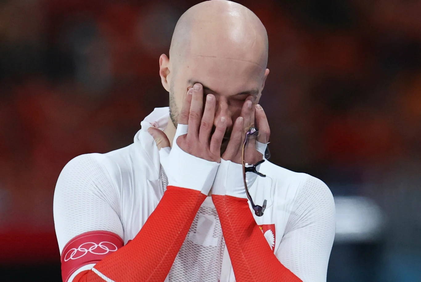 Piotr Michalski, zimowe igrzyska olimpijskie Mediolan-Cortina 2026 Sportowiec ubrany w biało-czerwony strój z emblematem olimpijskim na ramieniu, zakrywający twarz dłońmi w geście silnych emocji, stojący na tle rozmytego tłumu.