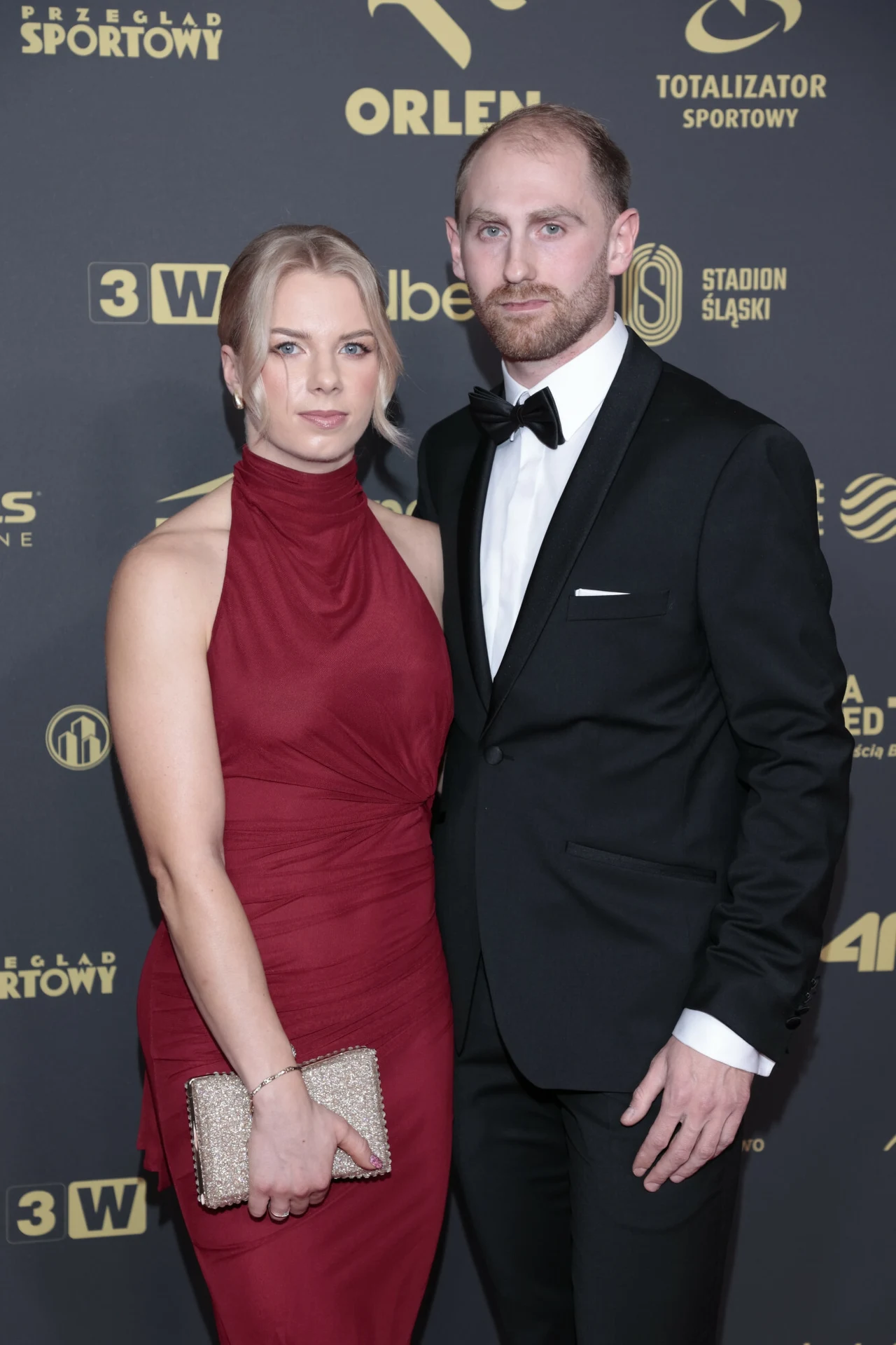 Elegancka kobieta w czerwonej sukni i mężczyzna w czarnym garniturze pozują razem na tle ściany z logotypami sponsorów podczas oficjalnego wydarzenia lub gali.