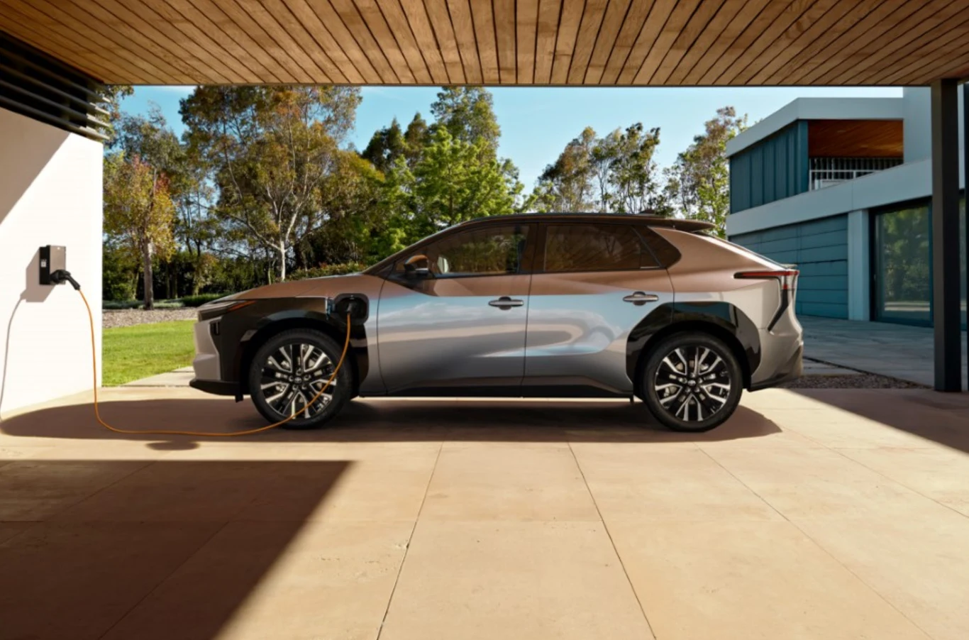 Toyota wprowadziła promocję na egzemplarze bZ4X z rocznika 2025. W wybranych wersjach Style i Executive rabat wynosi do 20 tys. zł. Samochód elektryczny w kolorze srebrno-brązowym ładuje się podłączony do domowej stacji ładowania, zaparkowany na nowoczesnym podjeździe przy budynku z przeszklonym tarasem, w tle widoczna zieleń ogrodu i nowoczesna architektura.