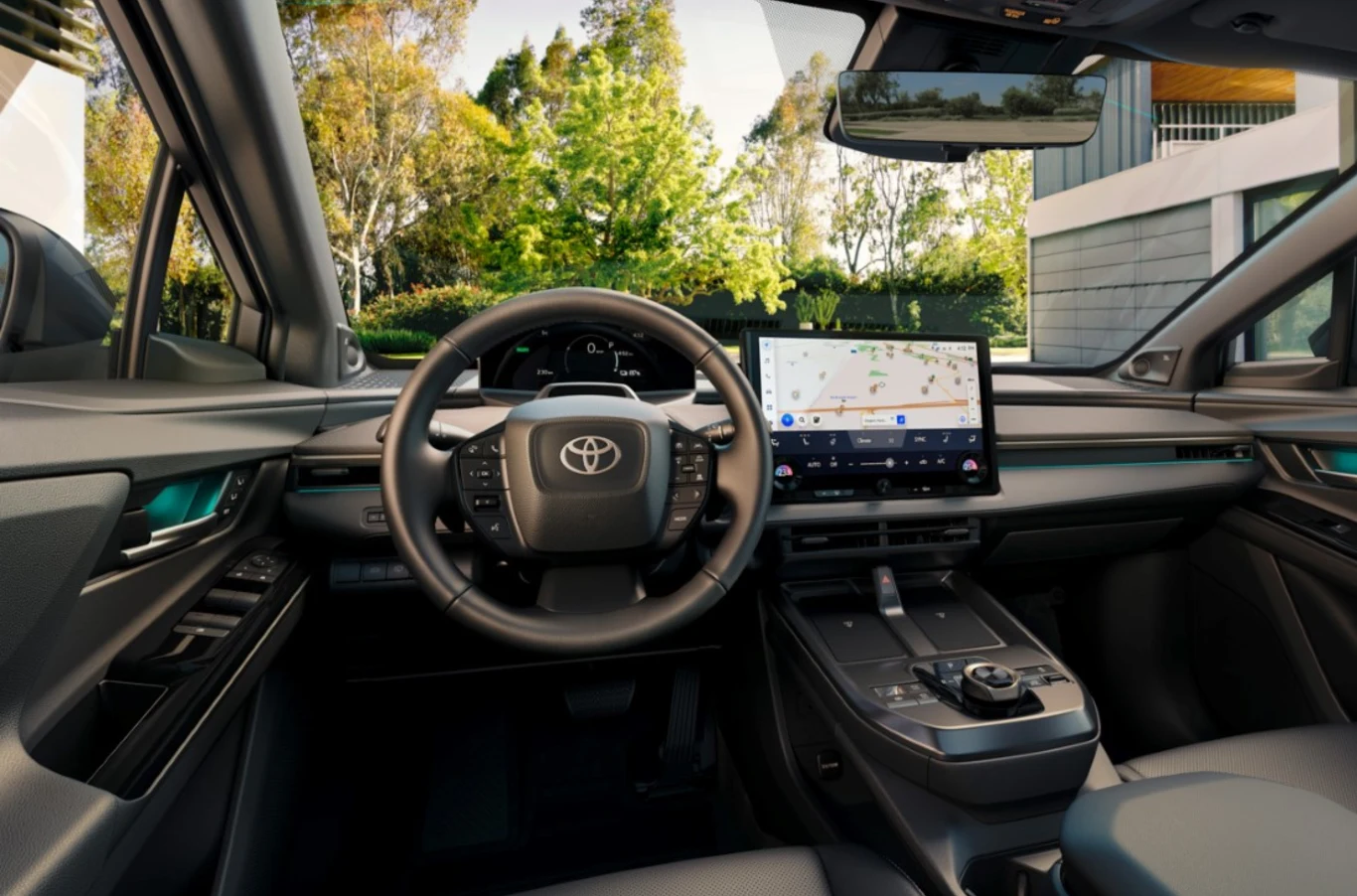 W salonach dostępne są również egzemplarze z rocznika 2026. Nowoczesna deska rozdzielcza samochodu marki Toyota z dużym ekranem nawigacji na środku, kierownica z logo firmy, widok na przejrzyste wnętrze i fragment ogrodu za szybą.