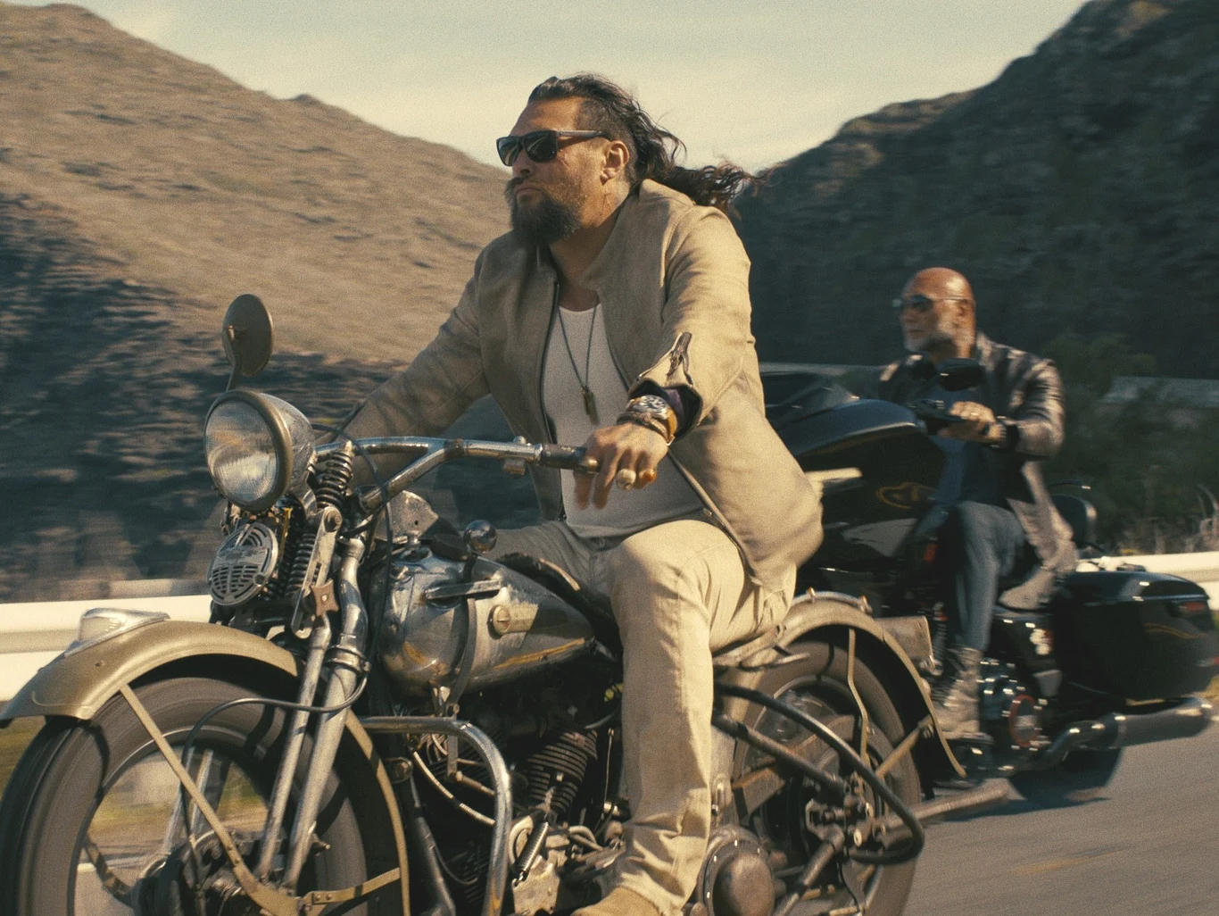 Jason Momoa i Dave Bautista w filmie "Ekipa wyburzeniowa" Mężczyzna z długimi włosami i brodą jedzie motocyklem po asfaltowej drodze przez górzysty teren. W tle drugi motocyklista na czarnym motocyklu, obaj w okularach przeciwsłonecznych, otoczonych pustynnym krajobrazem i jasnym światłem słonecznym.