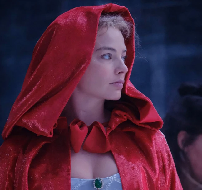 Margot Robbie w filmie "Wichrowe wzgórza" Kobieta o jasnej cerze ubrana w czerwony, aksamitny płaszcz z kapturem, patrzy w bok z poważnym wyrazem twarzy. Na szyi ma elegancką białą suknię z zielonym kamieniem, w tle rozmyta postać.