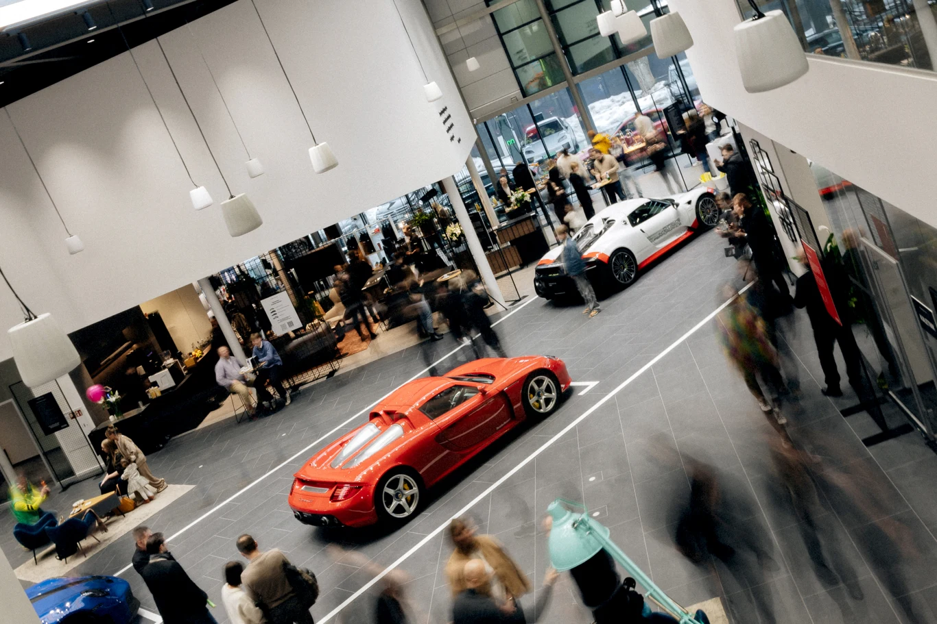 Luksusowe samochody sportowe eksponowane w nowoczesnym, przestronnym salonie otoczonym przez ludzi, którzy zwiedzają, rozmawiają i robią zdjęcia.