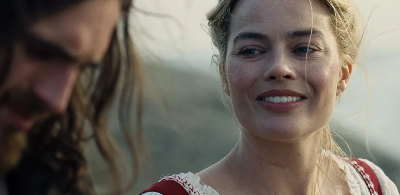 Kadr z filmu "Wichrowe wzgórza" Kobieta uśmiecha się delikatnie, spoglądając na mężczyznę z długimi włosami na tle rozmytego krajobrazu.