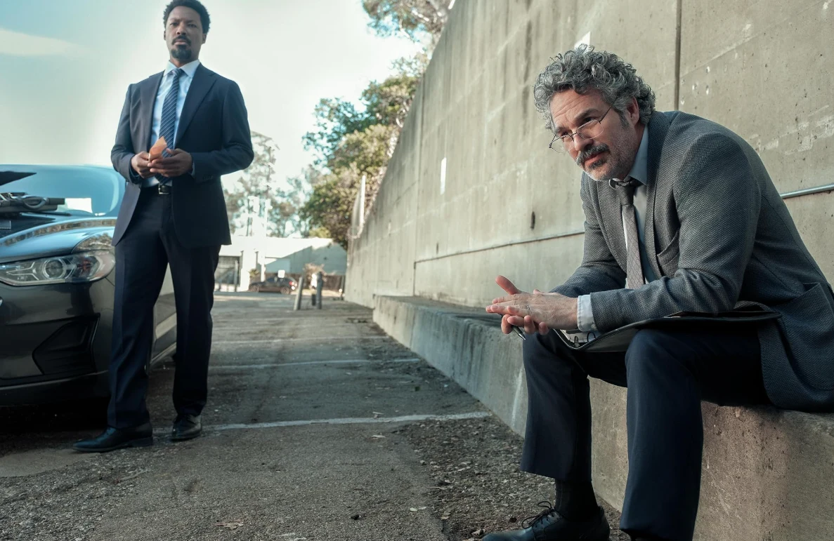 Mark Ruffalo i Corey Hawkins w filmie "Crime 101" Dwóch mężczyzn w garniturach znajduje się na parkingu przy betonowej ścianie. Jeden z nich siedzi zamyślony na krawędzi murka, drugi stoi obok sportowego samochodu. Scena wydaje się być napięta i poważna.