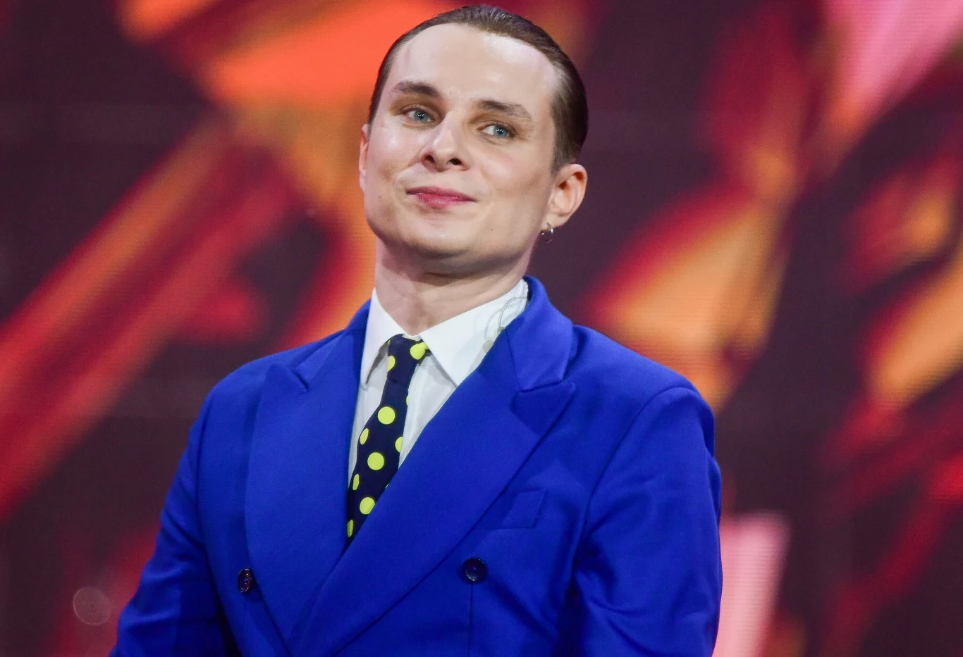 Ralph Kamiński zdradził datę premiery nowego albumu Mężczyzna w granatowym garniturze z białą koszulą i krawatem w żółte kropki, stojący na tle wielokolorowych, abstrakcyjnych wzorów.