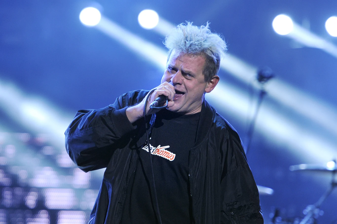 Kazik jest wieloletnim wokalistą Kultu Mężczyzna z jasnymi, rozczochranymi włosami śpiewa do mikrofonu na scenie koncertowej, ubrany w czarną kurtkę i koszulkę, w tle widoczne niebieskie światła sceniczne.