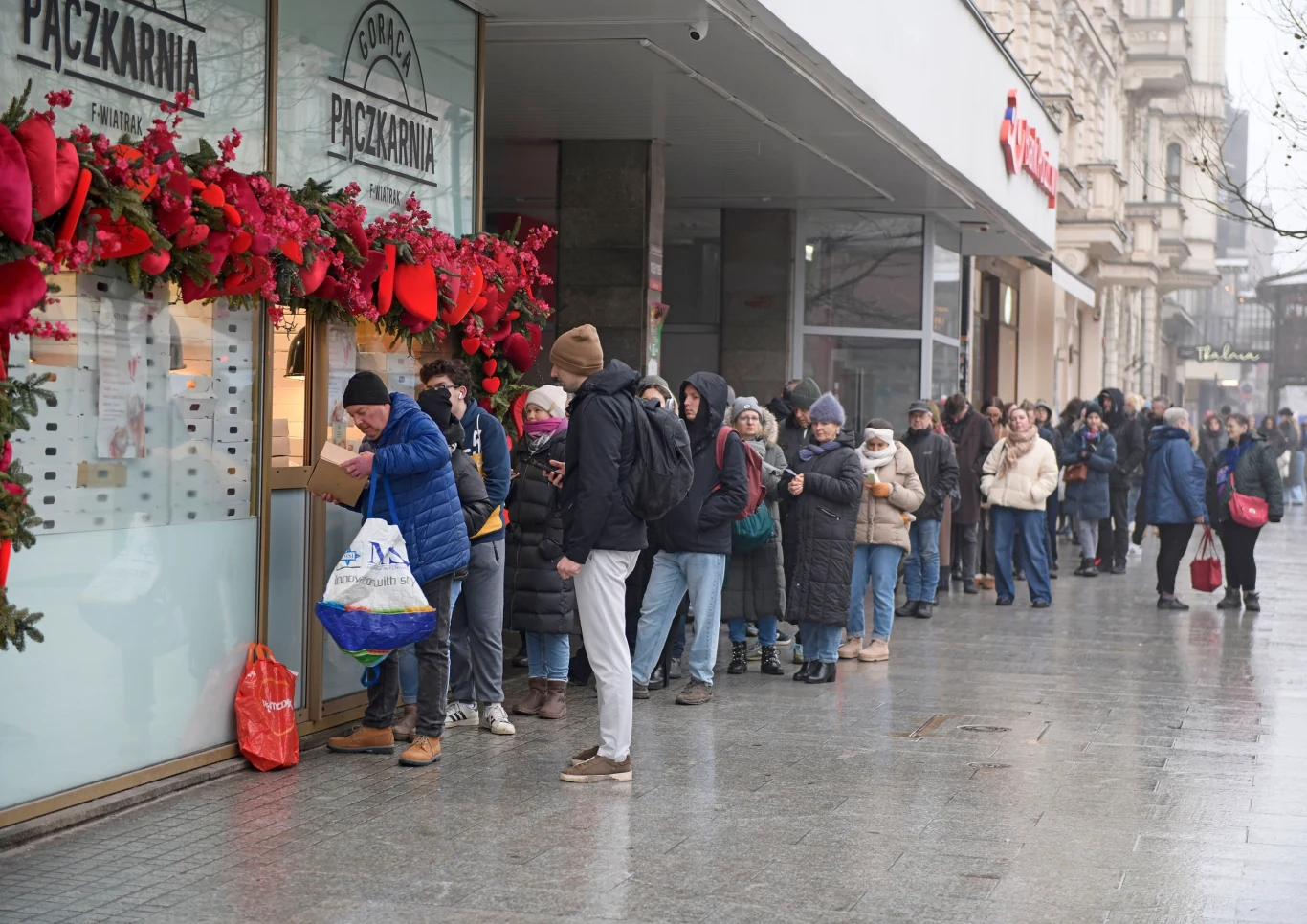 Długa kolejka ludzi ustawiona na chodniku przed pączkarnią, większość osób ubrana w zimowe kurtki i czapki, na wystawie lokalu świąteczne dekoracje oraz duży napis 'Pączkarnia', ulica miasta w tle.