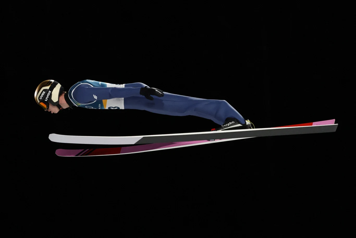 Skoczek narciarski w locie na tle czarnego tła, ubrany w kombinezon sportowy, z kaskiem i goglami, w aerodynamicznej pozycji na nartach.