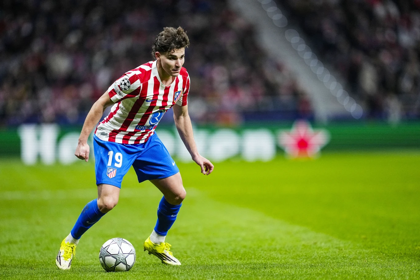 Piłkarz w stroju Atlético Madryt kontroluje piłkę podczas meczu na stadionie, otoczony przez rozmazane tło trybun z kibicami.